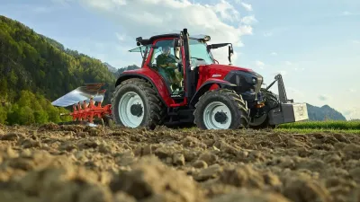 Červený traktor Lindner s pluhem pracuje na poli před horami.