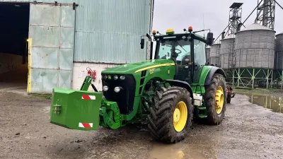 Zelený traktor John Deere před hospodářskou budovou.