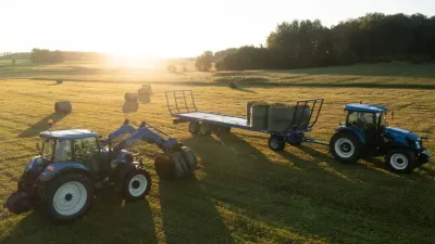 Dva modré traktory New Holland při manipulaci s balíky na louce.