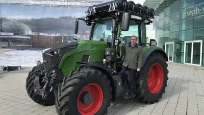 Zelený traktor Fendt s mužem před budovou.