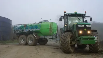 Zelený traktor John Deere táhne zelenou dvounápravovou cisternu Bauer na dvoře.