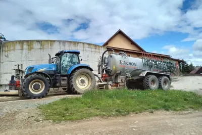 Modrý traktor táhne cisternu Pichon u zemědělské budovy