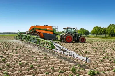 Traktor táhne postřikovač Amazone s rozloženými rameny na poli.