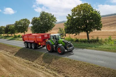 Traktor Fendt 700 Vario s červeným přívěsem jede po silnici.