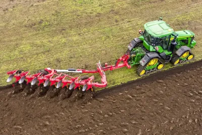 Pásový traktor táhne červený pluh Pöttinger při orbě.